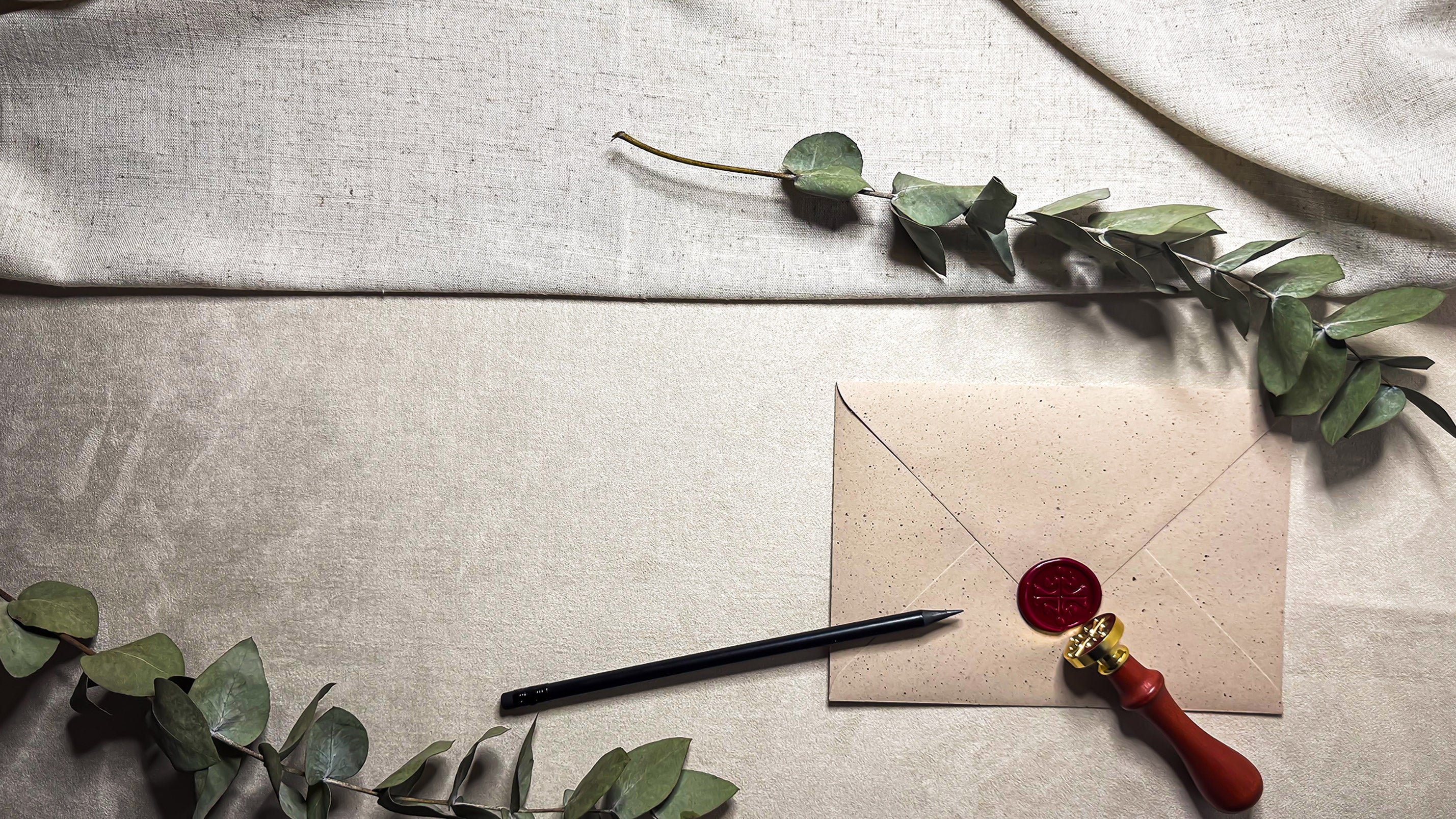 Horizontalna kompozicija pisemskega seta Letera z velikim prostorom za besedilo na levi. V desnem kotu je kraft ovojnica z rdečim voščenim pečatom, pečatnik in svinčnik, okvirjeno z vejami evkaliptusa.